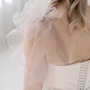 Puede incluir: Un vestido de novia blanco con falda de tul y un corpi&ntilde;o ajustado. El corpi&ntilde;o tiene una espalda con cordones y una delicada superposici&oacute;n de tul en un hombro.