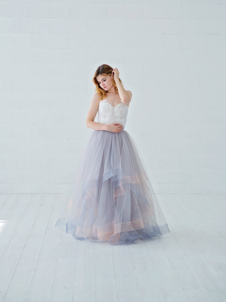 May include: A woman wearing a white lace bodice and a layered tulle skirt in shades of gray and pink. The skirt has a ruffled hem and is flowing.