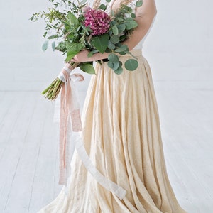 May include: A woman wearing a white top and a long, flowing, cream-colored skirt. She holds a bouquet of greenery and pink flowers tied with ribbons. The bouquet includes eucalyptus and a large pink hydrangea. The skirt has a subtle texture.