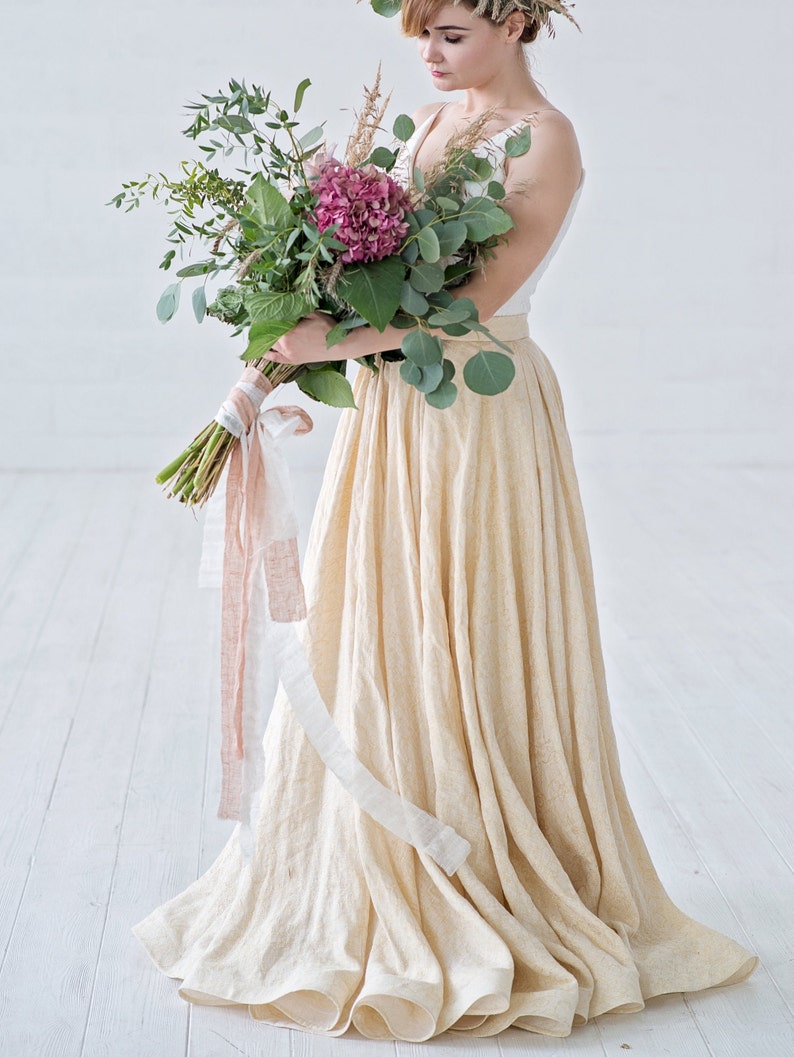 May include: A woman wearing a white top and a long, flowing, cream-colored skirt. She holds a bouquet of greenery and pink flowers tied with ribbons. The bouquet includes eucalyptus and a large pink hydrangea. The skirt has a subtle texture.