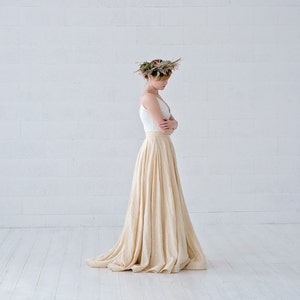 May include: A woman wearing a white top and a long, flowing beige skirt. She is also wearing a floral crown. The outfit is simple and elegant, with a focus on natural materials and a flowing silhouette. The skirt appears to be made of a soft, textured fabric.