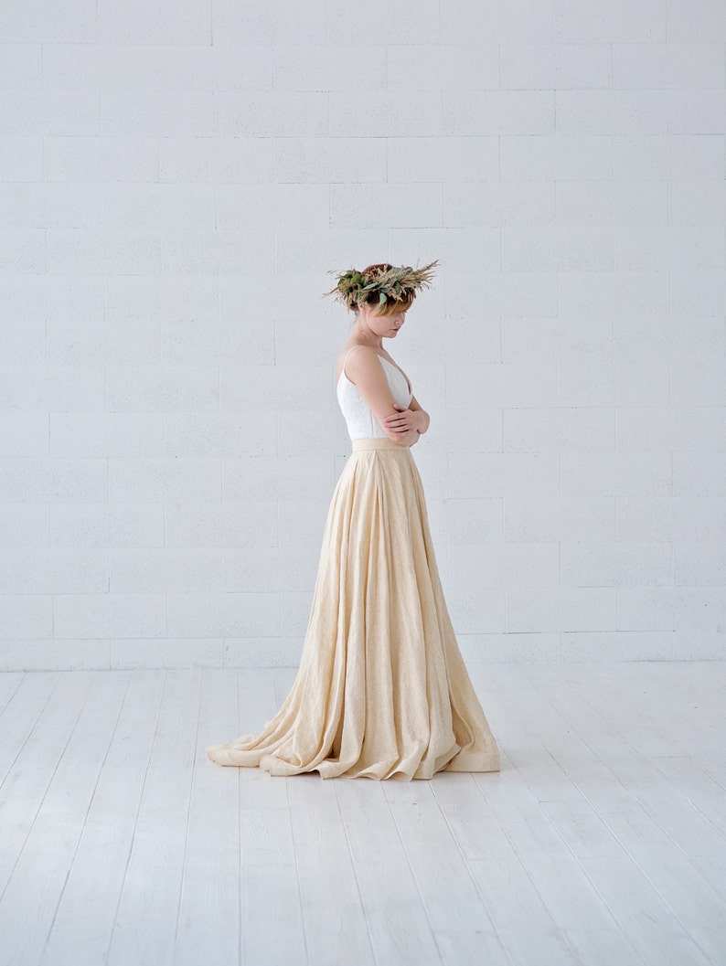 May include: A woman wearing a white top and a long, flowing beige skirt. She is also wearing a floral crown. The outfit is simple and elegant, with a focus on natural materials and a flowing silhouette. The skirt appears to be made of a soft, textured fabric.
