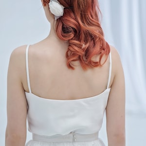 May include: A woman wearing a white satin top and a white lace skirt. Her hair is styled in loose curls and she has a white flower in her hair.