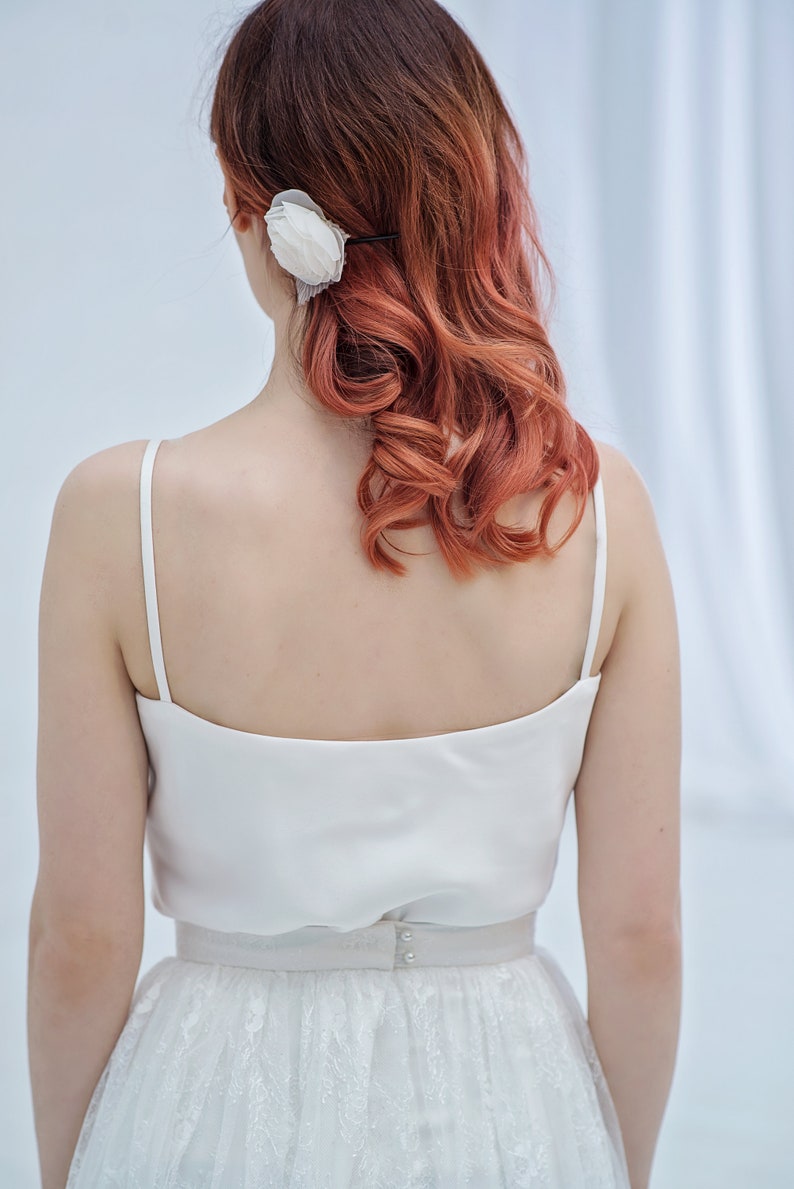 May include: A woman wearing a white satin top and a white lace skirt. Her hair is styled in loose curls and she has a white flower in her hair.