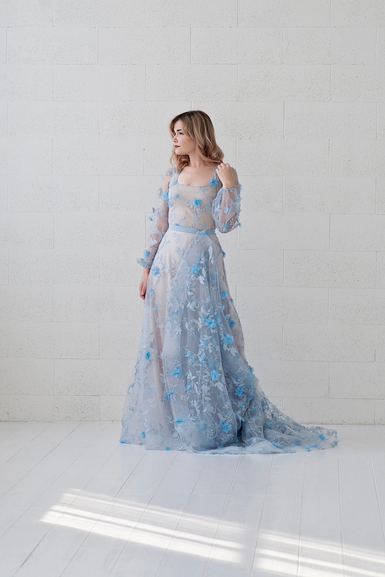 May include: A woman wearing a light blue gown with floral embroidery. The gown has long sleeves and a flowing skirt. The woman is standing in front of a white brick wall.