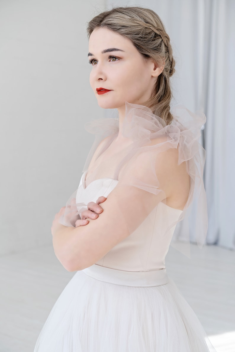 Puede incluir: Una mujer con un vestido de novia blanco con falda de tul y una superposici&oacute;n de tul transparente de color rosa claro en un hombro. El vestido tiene un escote de coraz&oacute;n y un corpi&ntilde;o ajustado.
