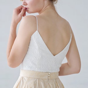 May include: A white lace top with spaghetti straps and a V-neckline is paired with a gold skirt. The skirt has a pleated design and a gold patterned belt. The back of the dress features three pearl buttons.