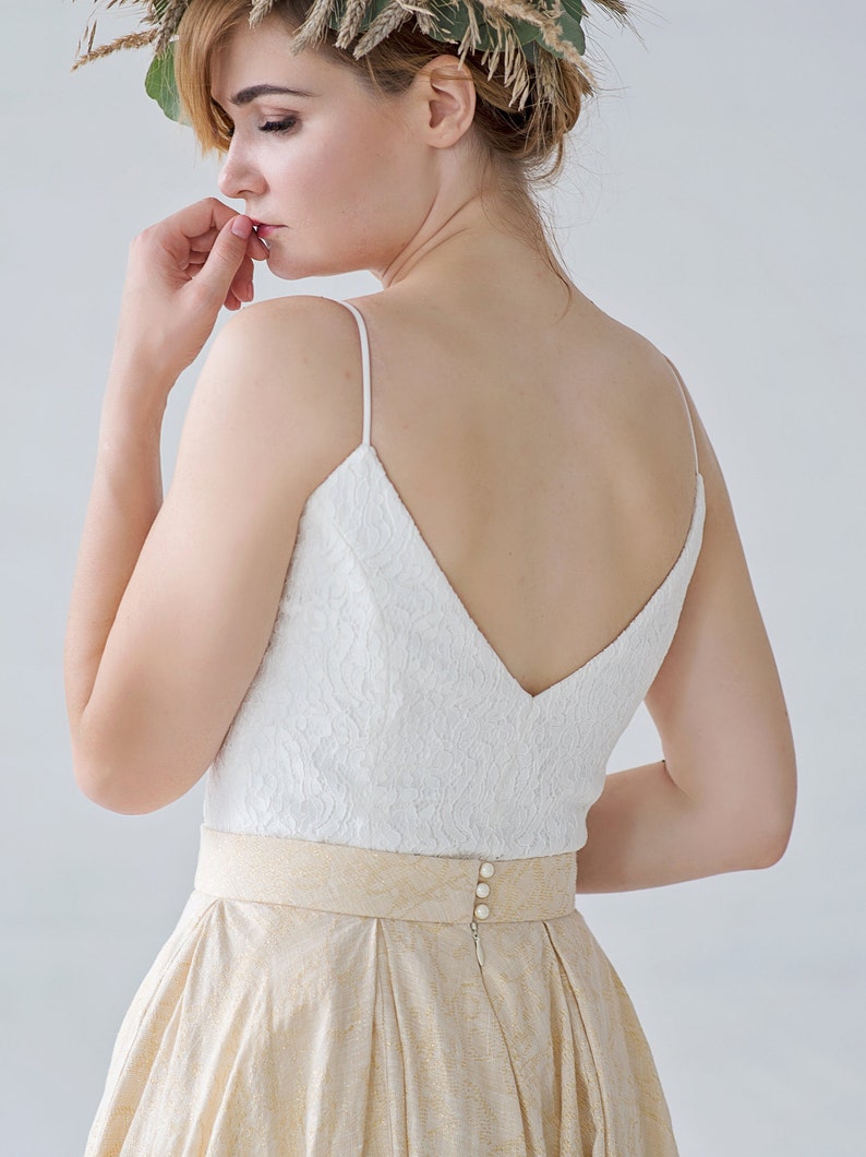 May include: A white lace top with spaghetti straps and a V-neckline is paired with a gold skirt. The skirt has a pleated design and a gold patterned belt. The back of the dress features three pearl buttons.