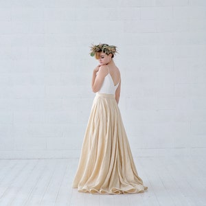 May include: A woman wearing a white spaghetti strap top and a long, flowing beige skirt. She is wearing a floral crown. The outfit is set against a white brick wall and white wooden floor.