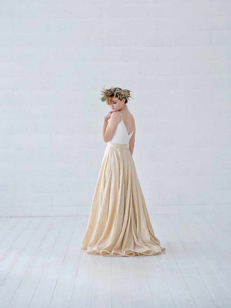 May include: A woman wearing a white spaghetti strap top and a long, flowing beige skirt. She is wearing a floral crown. The outfit is set against a white brick wall and white wooden floor.