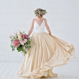 May include: A flowing, cream-colored skirt and a white top with a V-neck are featured in this image. The person is wearing a floral crown and holding a bouquet of pink and white flowers. The skirt is made of a light, airy fabric.