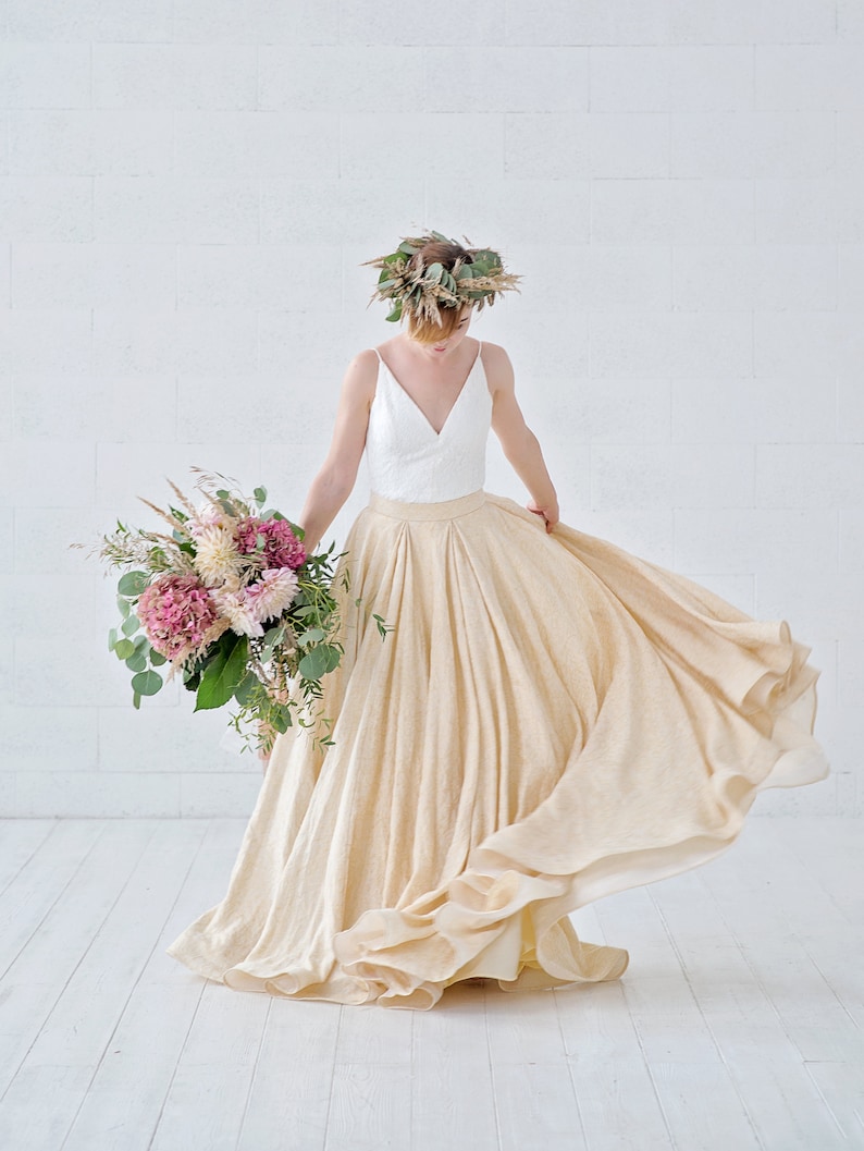 May include: A flowing, cream-colored skirt and a white top with a V-neck are featured in this image. The person is wearing a floral crown and holding a bouquet of pink and white flowers. The skirt is made of a light, airy fabric.