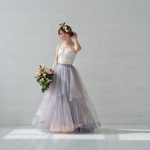 May include: A woman wearing a white lace bodice and a flowing gray tulle skirt with a floral crown. She is holding a bouquet of pink and white flowers.
