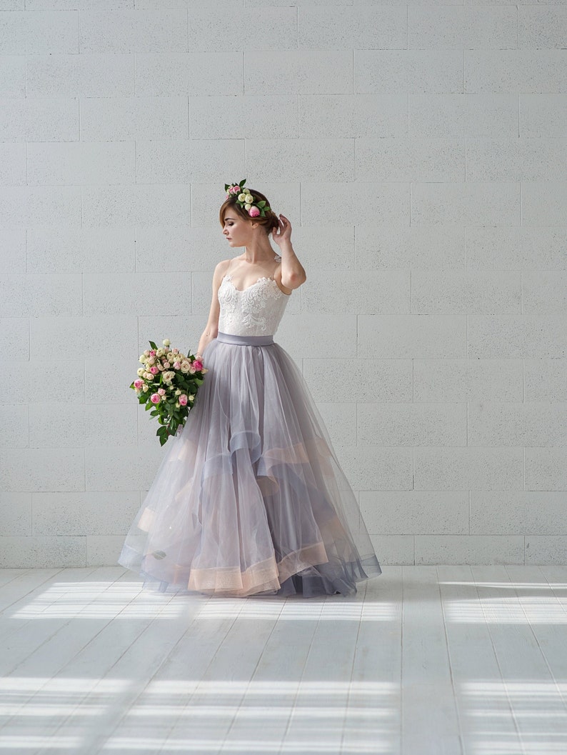 May include: A woman wearing a white lace bodice and a flowing gray tulle skirt with a floral crown. She is holding a bouquet of pink and white flowers.