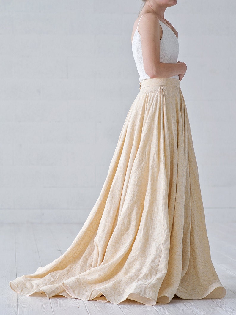 May include: A cream-colored, floor-length skirt with a subtle floral pattern. The skirt is paired with a white, textured camisole top. The skirt has a high waist and a flowing silhouette, suitable for formal occasions.