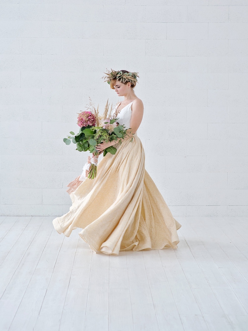 May include: A woman in a white top and a flowing, cream-colored skirt, holding a bouquet of flowers. She wears a floral crown. The background is a white brick wall and white wooden floor.