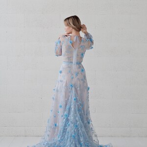 May include: A woman wearing a light blue gown with floral embroidery. The gown has a long train and a fitted bodice. The woman is standing in front of a white brick wall.