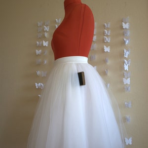 May include: A white tulle skirt with a satin waistband. The skirt is on a mannequin and is being displayed against a wall with white paper butterflies.