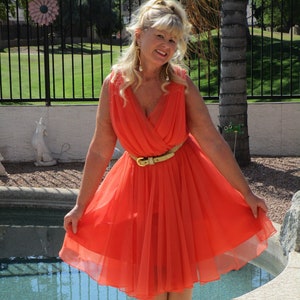 May include: A woman wearing a bright orange, flowing dress with a gold belt. She is standing by a swimming pool and smiling.