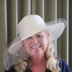Puede incluir: Un sombrero de paja blanco con ala ancha y un velo delicado. El sombrero tiene una forma clásica y está adornado con una fina red que cae sobre la cara. Un accesorio elegante.
