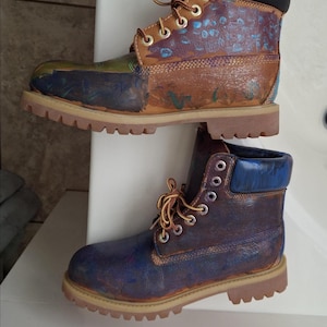May include: Two ankle boots with thick tan lug soles and lace-up closures. The top boot is tan with blue and green painted accents and a black padded collar. The bottom boot is dark blue and brown with a blue padded collar and silver eyelets.