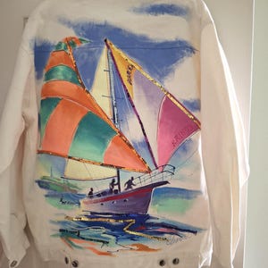 Può includere: Una giacca di jeans bianca con una scena dipinta a mano di una barca a vela sul retro. La barca ha vele colorate arancioni, verdi e rosa con paillettes dorate. Il testo KR1593 è visibile sulla vela. Lo sfondo raffigura un cielo azzurro.