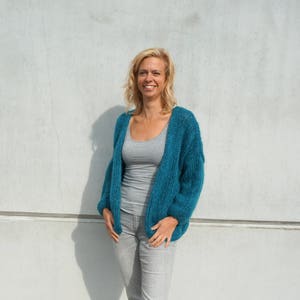 May include: A teal blue, open-front cardigan with long sleeves. The cardigan is made of a soft, textured material. The person in the image is wearing a gray tank top and gray jeans. The background is a light gray wall.