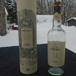 May include: A clear glass bottle of Oban 14 year old single malt Scotch whisky with a brown paper label. The bottle is sitting on a black surface next to a cardboard tube with a similar label. The tube has a picture of a rocky coastline with birds flying overhead.