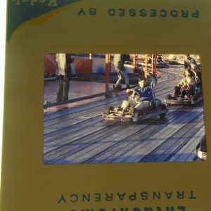May include: A slide showing a boy driving a go-kart on a wooden track. The boy is wearing a blue shirt and a blue hat. The track is surrounded by a metal fence. The slide is labeled 