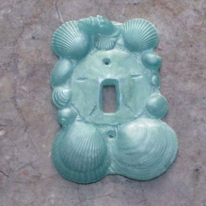 Puede incluir: Tapa de interruptor de luz de cerámica azul claro con un diseño de conchas marinas. La tapa presenta una variedad de conchas marinas, incluyendo vieiras, almejas y dólares de arena.