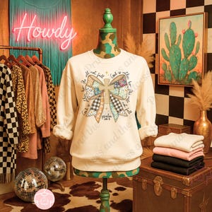May include: Cream-colored sweatshirt with a bow and cross design. The bow features a patchwork pattern with various colors and textures. Text on the sweatshirt reads "God is within her, she will not fall" and "Psalm 46:5". A rack of clothing and a neon "Howdy" sign are in the background.