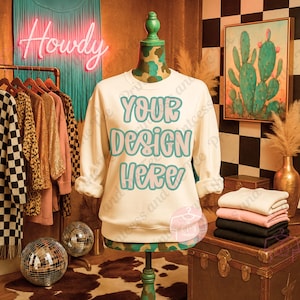 May include: A cream-colored sweatshirt with the text "YOUR DESIGN HERE" in turquoise lettering is displayed on a mannequin. The mannequin has a camouflage pattern on the neck and is set against a backdrop of clothing racks and decorative items.