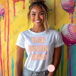 Puede incluir: Camiseta blanca con el texto "YOUR DESIGN HERE" en fuente de burbujas. El fondo presenta pelotas de béisbol con detalles en rosa y goteos de pintura. El tema general está relacionado con el diseño personalizado o la ropa personalizada.