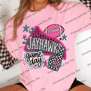 Op de afbeelding: Een lichtroze t-shirt met een grafische print met de tekst JAYHAWKS en game day. Het ontwerp bevat een football, een geblokte schuimvinger met het nummer 1, sterren en een wimpel met luipaardprint. De mouwen hebben een zwart-wit dambordpatroon.