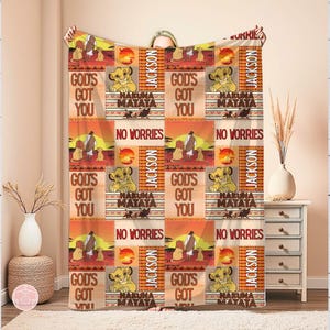 Puede incluir: Una manta beige y naranja con imágenes de dibujos animados de leones, los textos "GOD'S GOT YOU", "HAKUNA MATATA" y "NO WORRIES". El nombre "JACKSON" también está impreso en la manta. La manta se muestra en interiores.