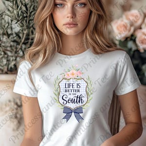 May include: White t-shirt featuring a floral and bow design with the text "Life is Better in the South." The design includes a blue and white gingham pattern and a navy blue bow. The shirt is a classic crew neck.