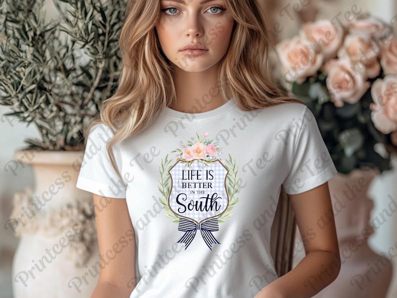 May include: White t-shirt featuring a floral and bow design with the text "Life is Better in the South." The design includes a blue and white gingham pattern and a navy blue bow. The shirt is a classic crew neck.