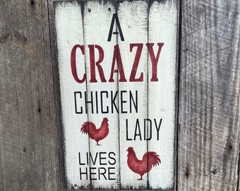 May include: A rectangular wooden sign with the text "A CRAZY CHICKEN LADY LIVES HERE" in black and red lettering. Two red rooster illustrations are featured. The sign is mounted on a weathered wooden wall.