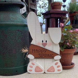 以下が含まれることがあります： 「Happy Spring」と書かれた茶色のニンジン型の看板を持った白い木製のウサギ。ウサギはピンク色の足と茶色の鼻をしています。