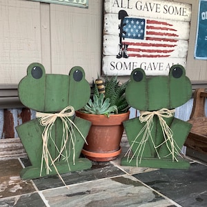 Puede incluir: Dos figuritas de rana de madera verde con una pajarita de paja. Las ranas están sentadas en un suelo de baldosas. El cartel detrás de las ranas dice "All Gave Some, Some Gave All".