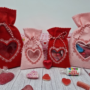 Op de afbeelding: Vier vilten Valentijnsdag-geschenkzakken met hartvormige ramen. De zakken zijn rood en roze en hebben een witte kantversiering. De zakken zijn gevuld met hartvormige snoepjes en andere lekkernijen. De zakken zijn gebonden met rood en wit touw.