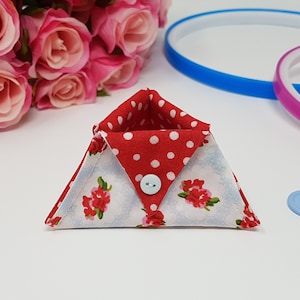 Puede incluir: Un pequeño contenedor de almacenamiento triangular de tela con un forro de lunares rojos y blancos y un exterior de estampado floral. El contenedor tiene un botón blanco en la parte delantera.