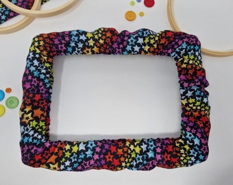 Puede incluir: Un aro de bordado negro con una cubierta de tela estampada con estrellas de colores. La tela es un arcoíris de colores con estrellas multicolores sobre un fondo negro.
