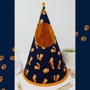 Puede incluir: Una cama para mascotas con forma de tipi colgante, azul y naranja, con estampado de zorro. La cama tiene una abertura redonda y un ribete amarillo.