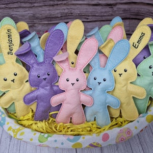Op de afbeelding: Een mand gevuld met pastelkleurige vilten konijnen speelgoed. De konijnen hebben geborduurde namen, waaronder "Benjamin" en "Emma".