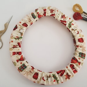 以下が含まれることがあります： クリスマスのテーマの白い布製のリース。リースはサンタクロース、雪だるま、プレゼント、星などの赤と緑の画像で飾られています。リースは、金色のハサミと糸巻き2つのある白い表面の上にあります。