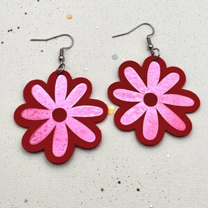 Peut inclure: Une paire de boucles d'oreilles en forme de fleur. Chaque boucle d'oreille présente un contour rouge avec un motif de fleur rose. Les boucles d'oreilles ont des crochets d'oreille argentés. Les boucles d'oreilles sont sur une surface blanche avec des paillettes.