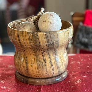 May include: A decorative wooden bowl with a unique grain pattern, holding two spherical ornaments and a small pine cone. The bowl is light brown with dark brown streaks and sits on a round base. The ornaments are white and silver.