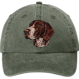 May include: A green, adjustable baseball cap featuring an embroidered image of a brown and white dog's head. The dog's fur is detailed with various shades of brown and white, and the cap has a curved brim and a top button.