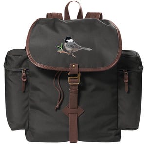 May include: A dark green canvas backpack with brown leather accents. It features a drawstring closure and a small embroidered image of a chickadee on a branch.